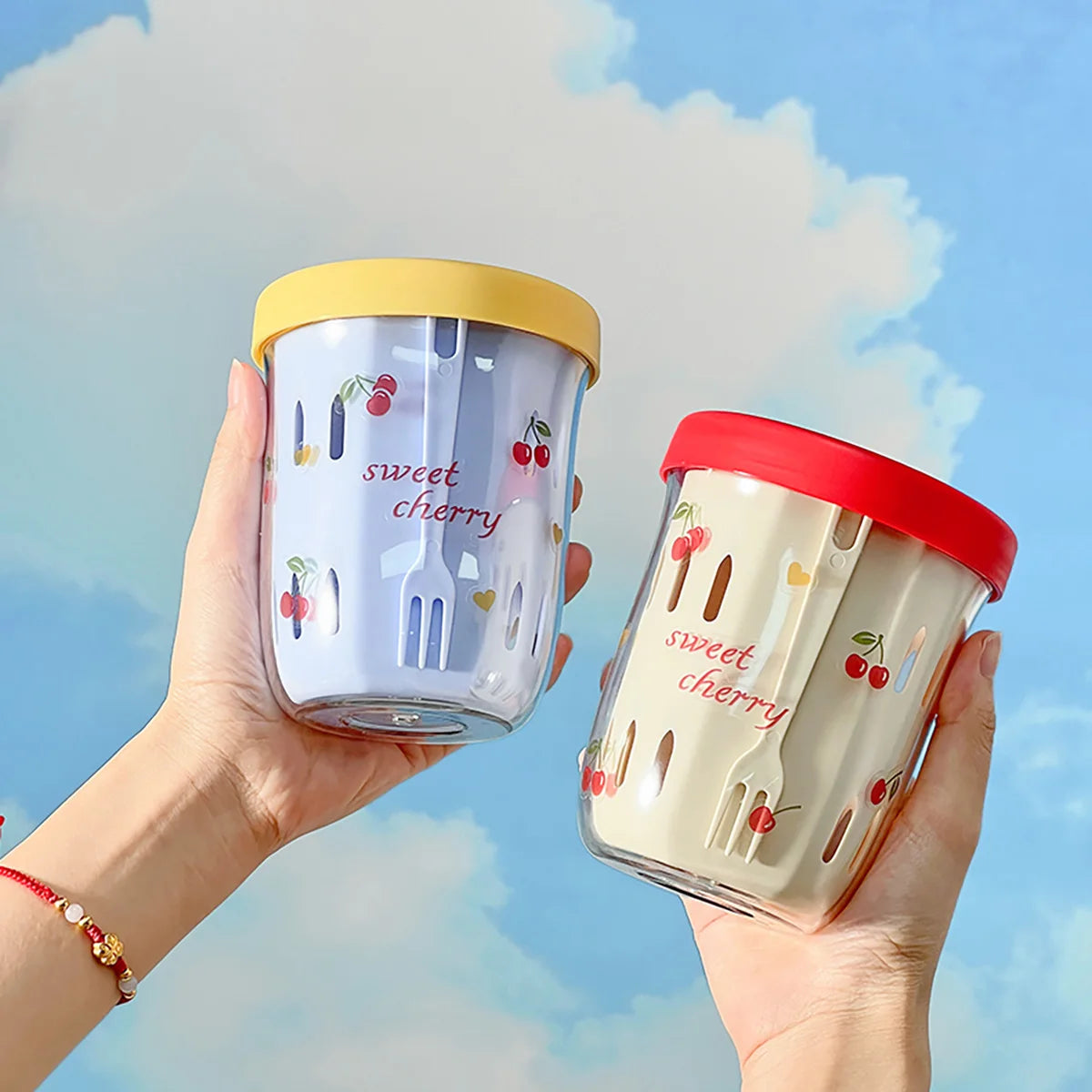 Doppelwandige Frühstücks- und Salatbecher mit Siebeinsatz in verschiedenen Farben – handgehalten vor blauem Himmel.